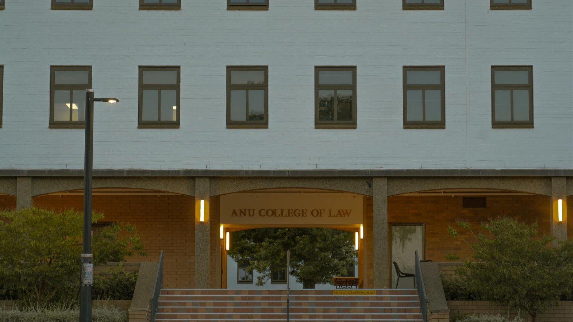 Front entrance of the ANU College of Law building at dusk, with warm lights illuminating the doorway and surrounding trees.