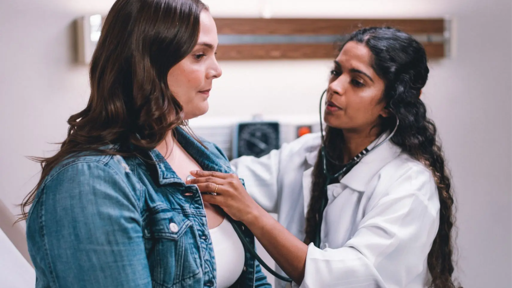 Doctor using a stethoscope to examine a patient in a clinic room.