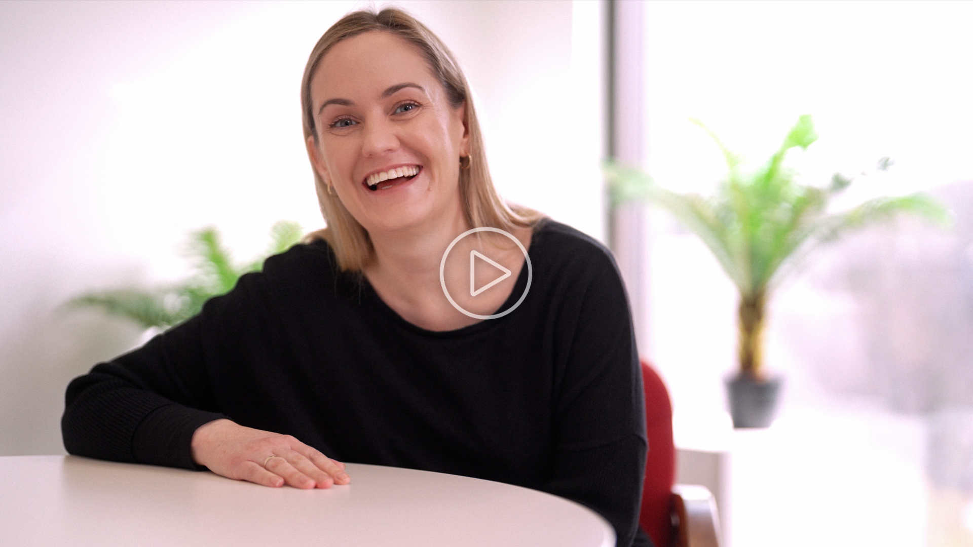 Person smiling while seated at a round table in a bright room with plants in the background.