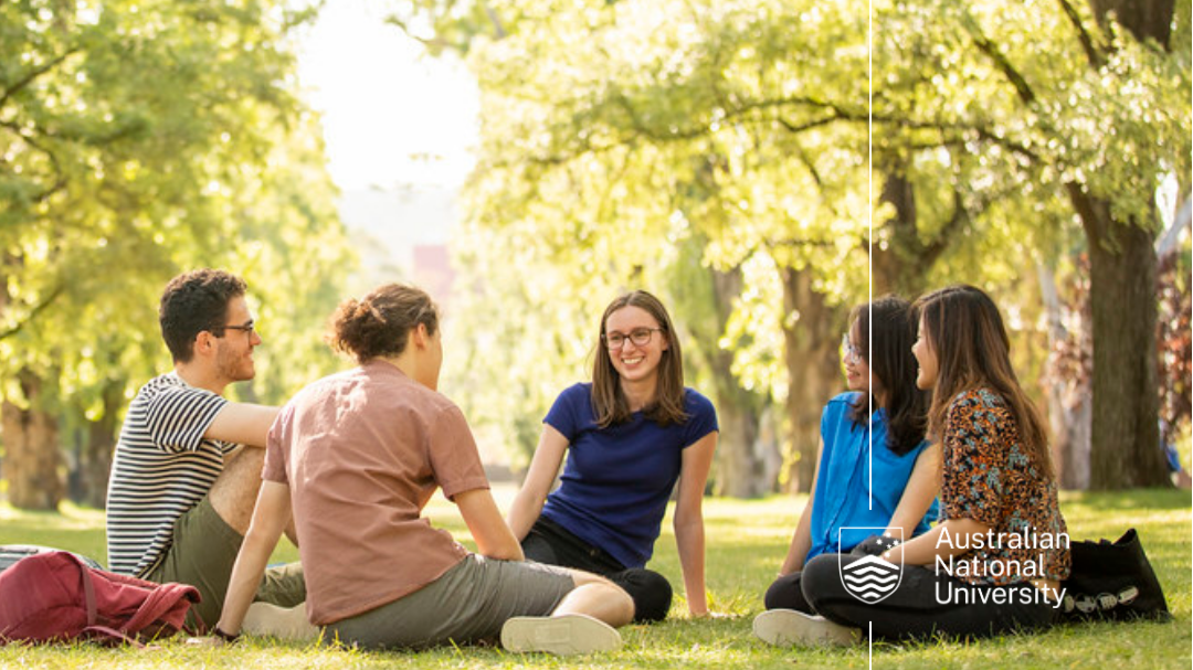 Five students sitting on grass under trees, chatting on campus.