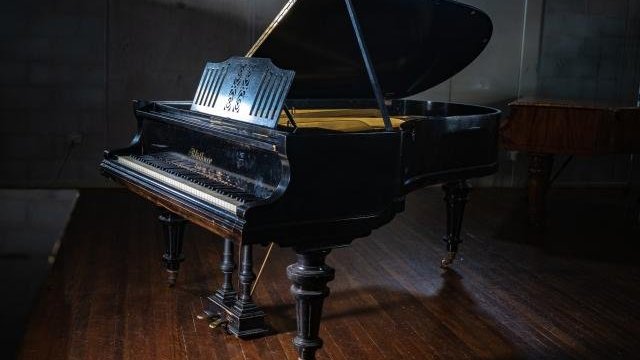 A black grand piano with an open lid sits on a polished wooden floor in a dimly lit room, light highlights the pianos glossy surface.