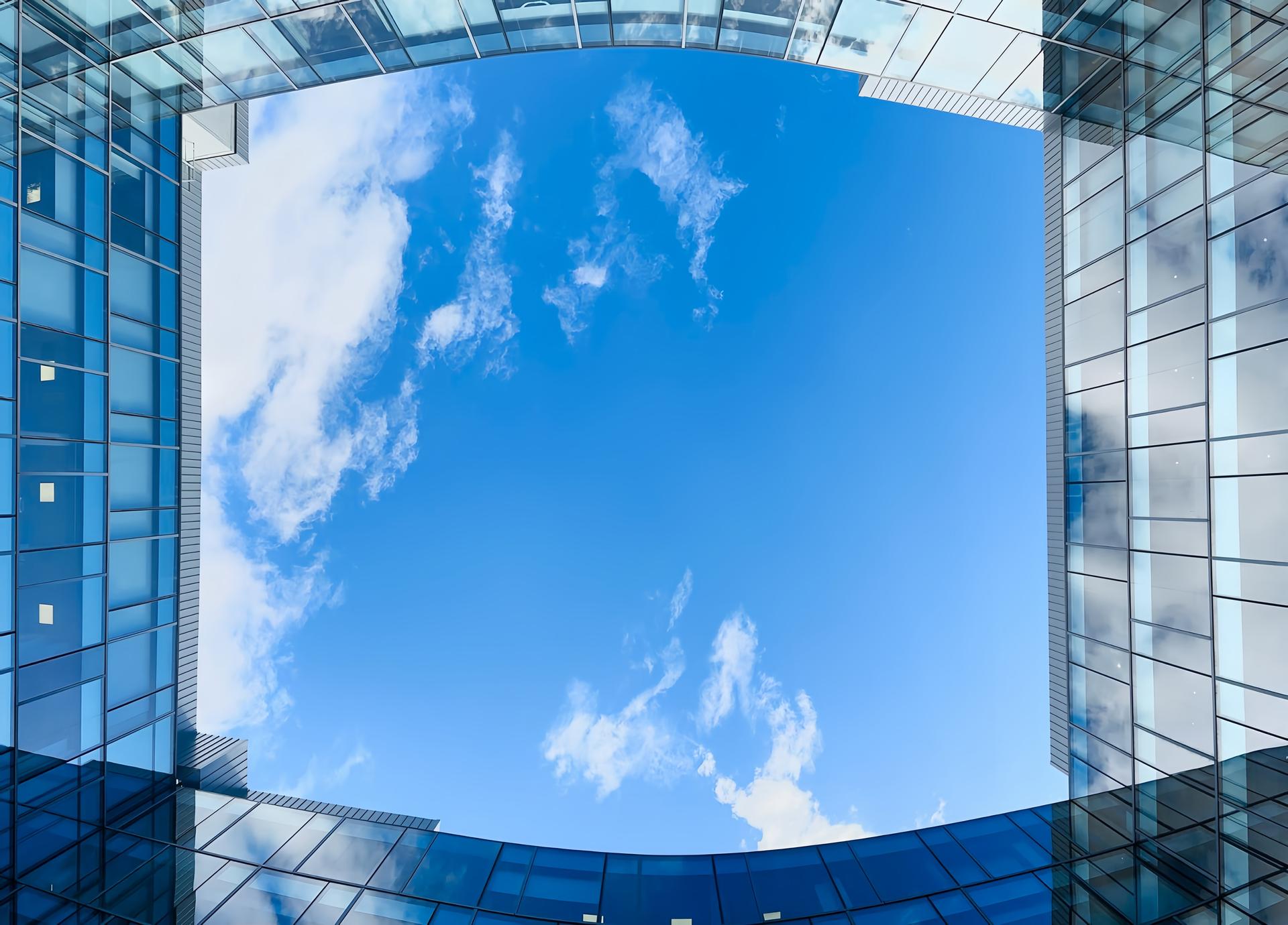 Open sky framed by the curved glass walls of a modern building, viewed from the courtyard looking straight up.