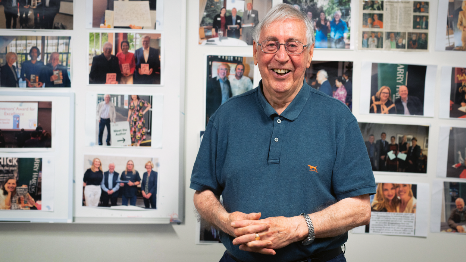 Emeritus Fellow, Colin Steele, smiling while standing in front of a wall covered with photos of events, people and achievements. 