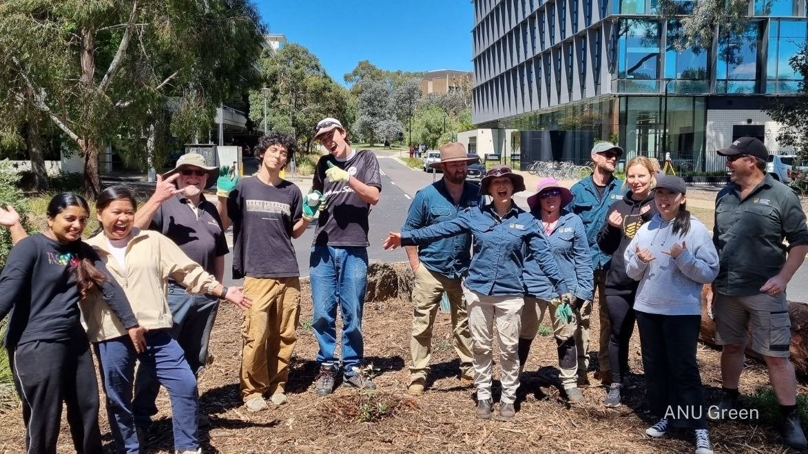 Group of students and staff smiling and posing during a campus sustainability activity, standing around a newly planted garden bed near an ��������building.
