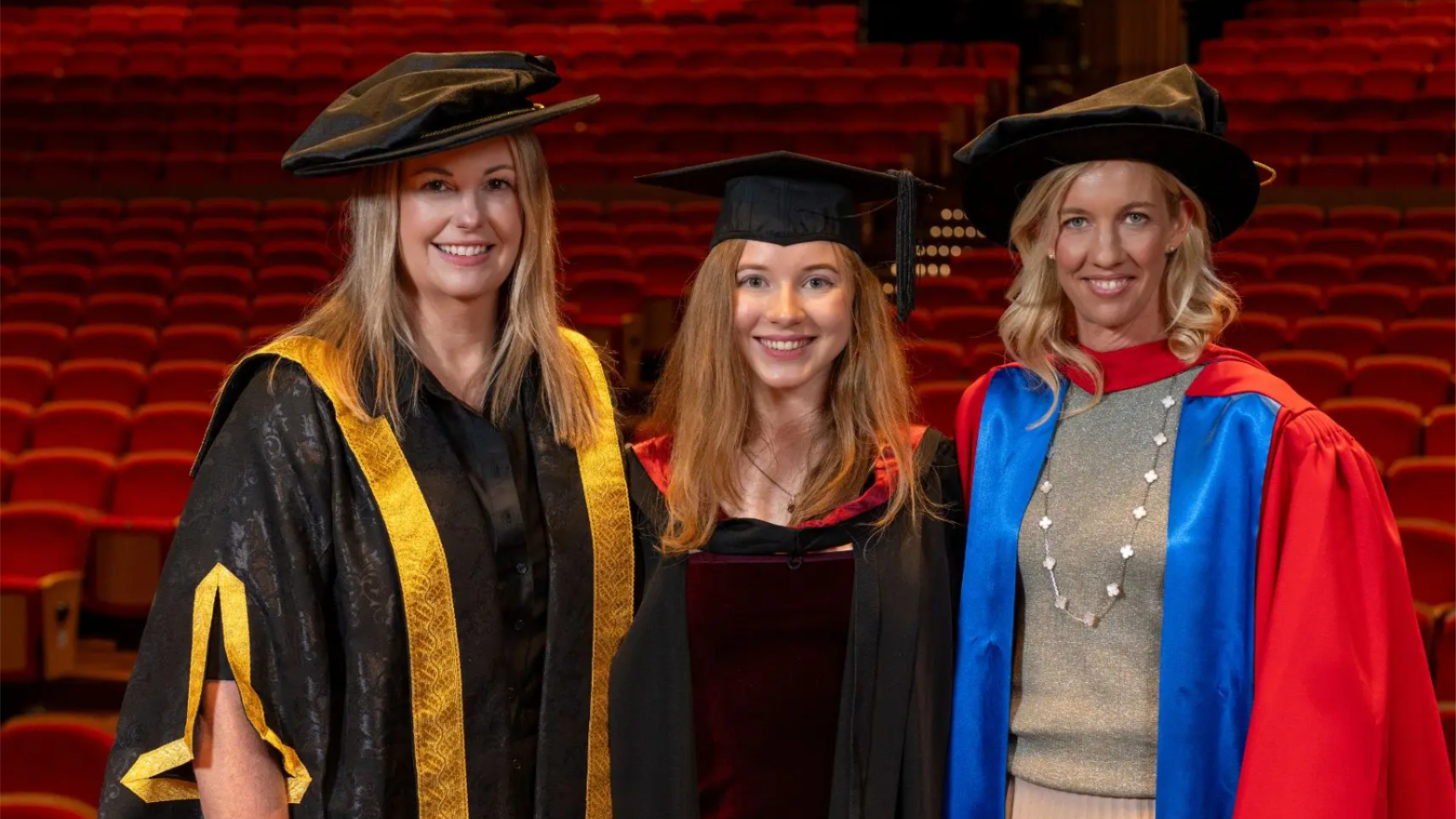Three women in academic regalia smiling in a theatre with red seats during a graduation ceremony.