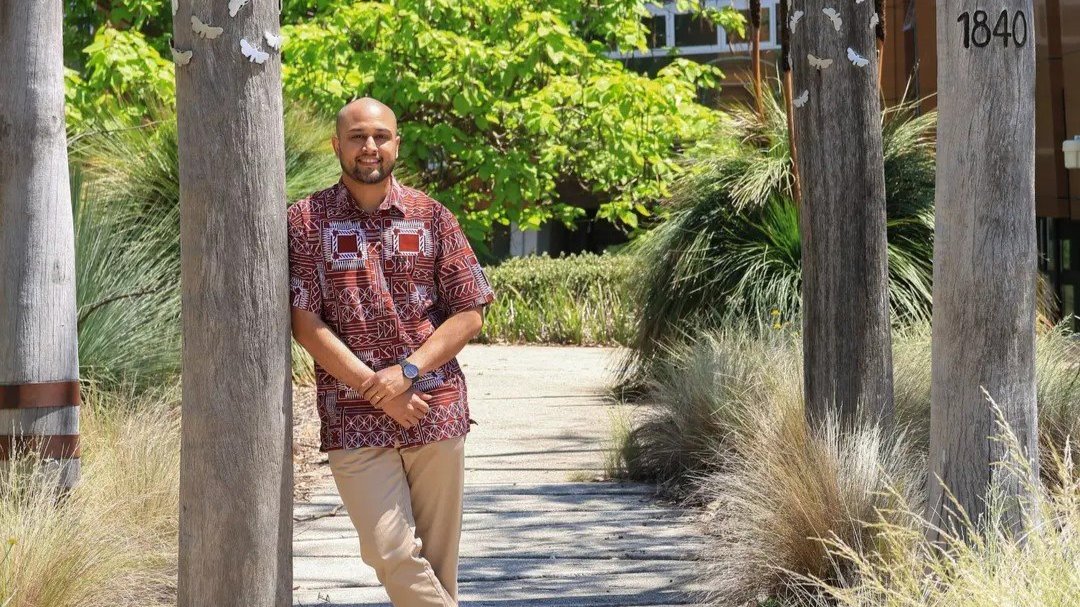 Person standing on a tree-lined campus pathway with landscaped greenery in the background.