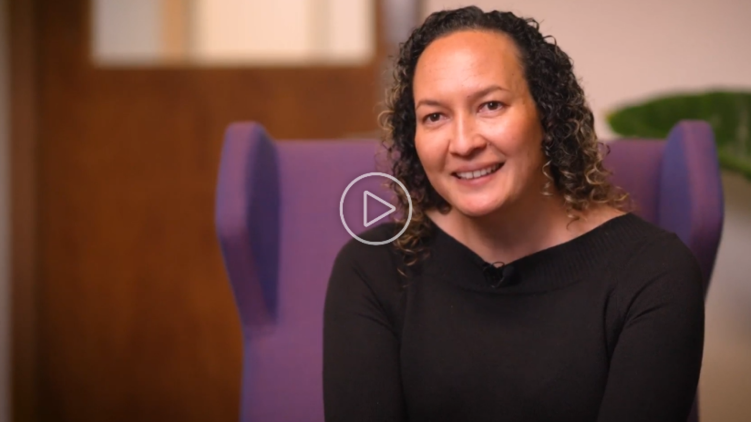 Fiona Cornforth smiling while seated on a purple chair, wearing a black top, with a blurred background featuring a wooden door and a green plant.