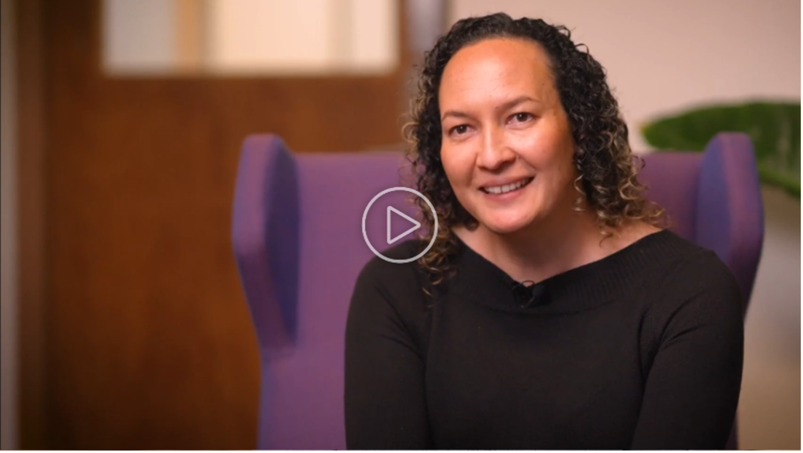 Fiona Cornforth smiling while seated on a purple chair, wearing a black top, with a blurred background featuring a wooden door and a green plant.
