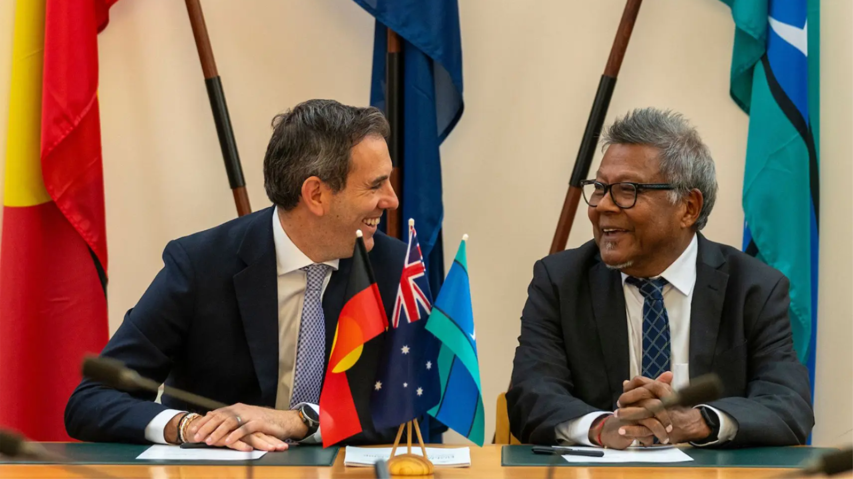 Treasurer Jim Chalmers and Professor Peter Yu smiling in conversation with flags the Aboriginal Flag, Australian flag and Torres Strait Island flags in the foreground and background. 