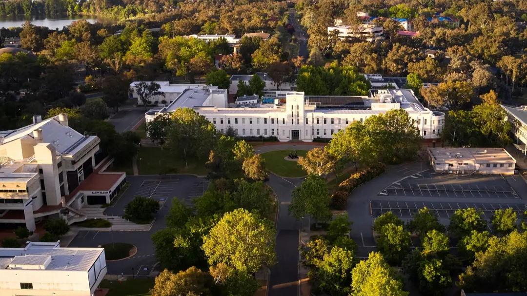 Aerial view of the Australian National University campus with the ANU School of Art and Design building centred, surrounded by trees, with Lake Burley Griffin and Canberra’s hills visible in the background. 