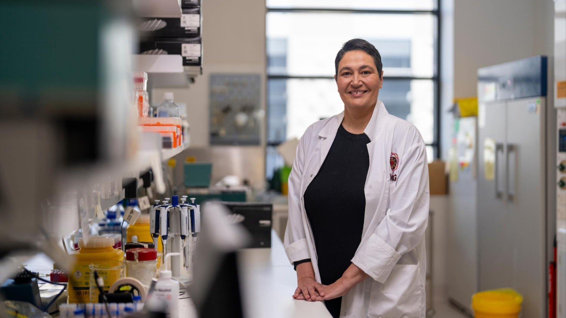 A scientist in a white lab coat smiles while standing at a lab bench surrounded by equipment, with bright natural light from a large window behind them.