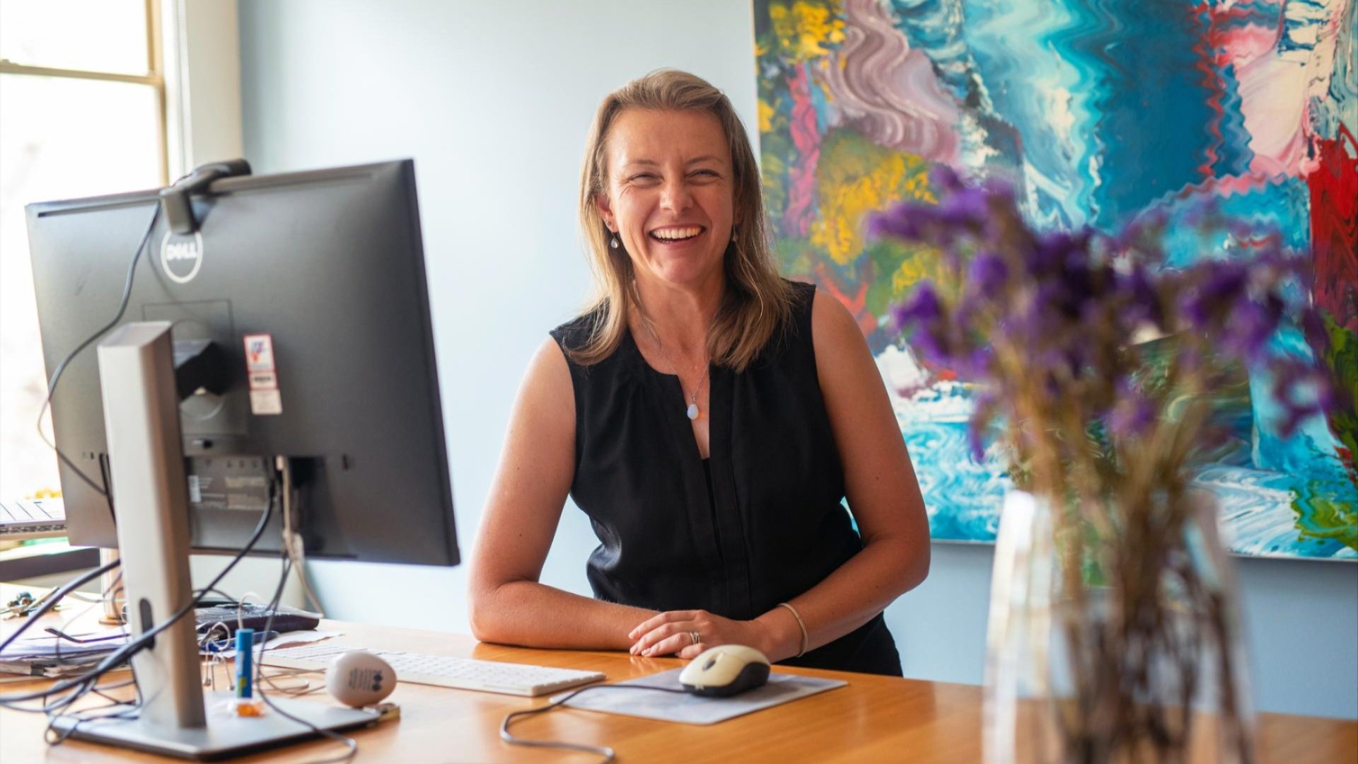 Smiling woman seated at a desk with a computer, abstract artwork on the wall behind her.