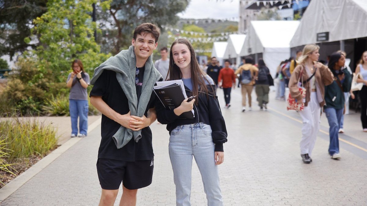 Two students smiling on a campus walkway, one holding a folder, with other students and white tents in the background.