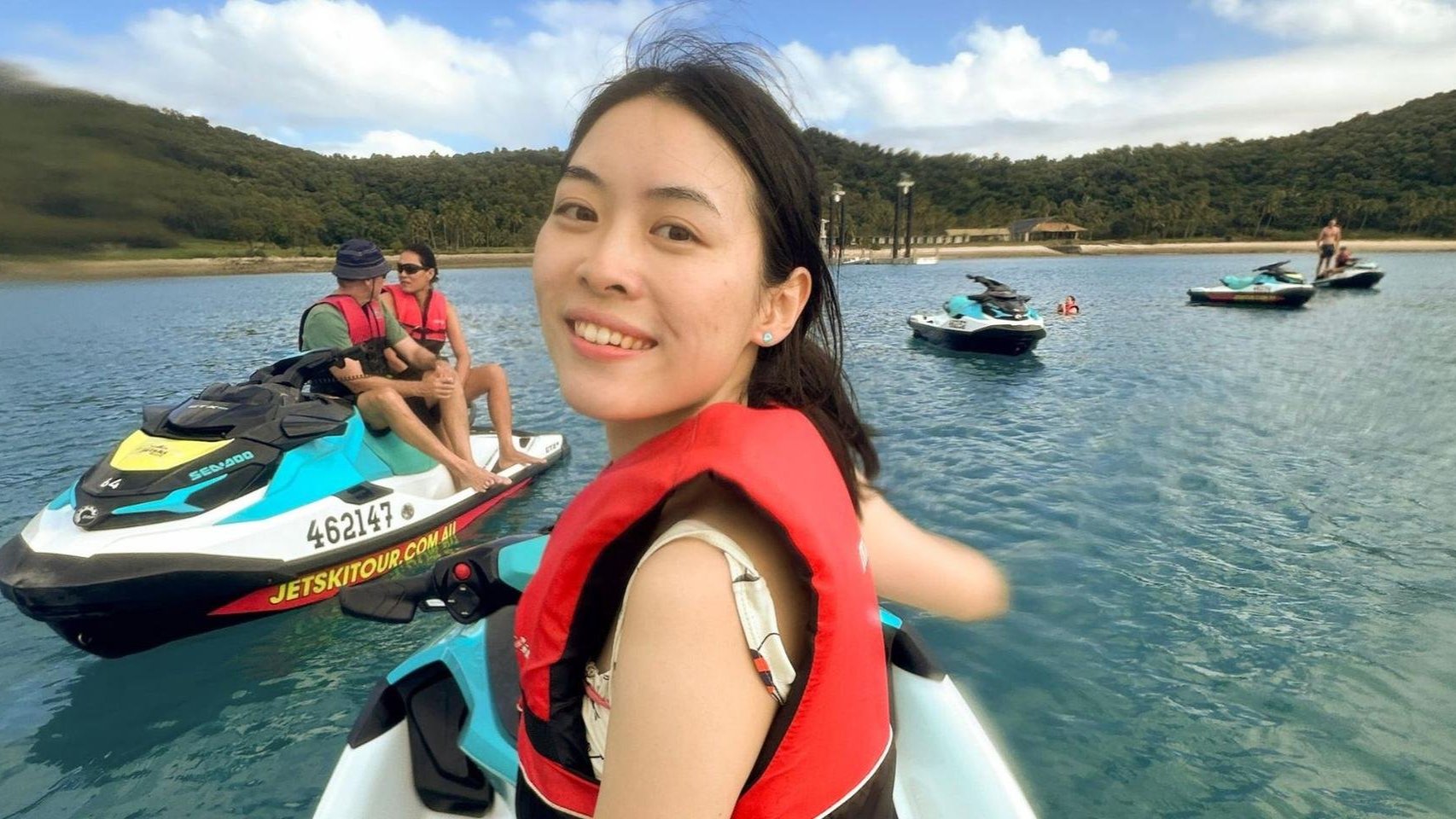 A woman wearing a red life jacket smiles at the camera while sitting on a jet ski in clear blue water, with other people on jet skis and green hills in the background under a partly cloudy sky.