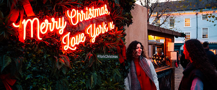 Two students at York Christmas Market stand in front of an illuminated sign reading ''Merry Christmas Love York''