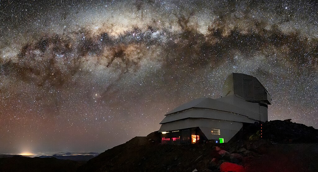 This image captures not only Vera C. Rubin Observatory, a Program of NSF’s NOIRLab, but one of the celestial specimens Rubin Observatory will observe when it comes online: the Milky Way. The bright halo of gas and stars on the left side of the image highlights the very center of the Milky Way galaxy. The dark path that cuts through this center is known as the Great Rift, because it gives the appearance that the Milky Way has been split in half, right through its center and along its radial arms. In fact, the Great Rift is caused by a shroud of dust, which blocks and scatters visible light. This dust makes the Great Rift a difficult space to observe. Fortunately, Rubin is being built to conduct the Legacy Survey of Space and Time (LSST). This survey will observe the entire visible southern sky every few nights over the course of a decade, capturing about 1000 images of the sky every night and giving us a new view of our evolving Universe. The LSST is set to probe four areas of science, one of which is mapping the Milky Way to answer questions about its structure and formation. Once Rubin Observatory comes online in 2024, scientists will be able to access the data taken every night of operation. Rubin Observatory is a joint initiative of the National Science Foundation and the Department of Energy (DOE). Once completed, Rubin will be operated jointly by NSF’s NOIRLab and DOE's SLAC National Accelerator Laboratory to carry out the Legacy Survey of Space and Time. Español: Esta imagen no sólo captura el Observatorio Vera C. Rubin, un Programa de NOIRLab de NSF, sino que también a uno de los especímenes celestes que el Observatorio Rubin observará cuando entre en funcionamiento: la Vía Láctea. El halo brillante de gas y estrellas en la parte izquierda de la imagen destaca el centro mismo de nuestra galaxia. El sendero oscuro que atraviesa este centro se conoce como la Gran Grieta, porque da la impresión de que la Vía Láctea estuviese partida en dos, justo a través de su centro y a lo largo de sus brazos espirales. En realidad, la Gran Grieta está formada por un manto de polvo que bloquea y dispersa la luz visible, convirtiéndola en un espacio difícil de observar. Por suerte, Rubin se está construyendo para llevar a cabo la Investigación del Espacio-Tiempo como Legado para la Posteridad (LSST por sus siglas en inglés). Esta investigación observará por una década todo el cielo austral visible, capturando unas 1.000 imágenes del cielo cada noche y ofreciéndonos una nueva perspectiva de nuestro Universo en evolución. El LSST está destinado a explorar cuatro áreas de la ciencia, y una de ellas es mapear la Vía Láctea para responder interrogantes sobre su estructura y formación. Una vez que el Observatorio Rubin entre en operación en 2024, los científicos podrán acceder a los datos recopilados en cada noche de operación. El Observatorio Rubin es una iniciativa conjunta de la Fundación Nacional de Ciencias y el Departamento de Energía (DOE). Una vez completado, el Observatorio Rubin será operado conjuntamente por NOIRLab de NSF y el Laboratorio Nacional del Acelerador SLAC de DOE para llevar a cabo el estudio denominado Legacy Survey of Space and Time.