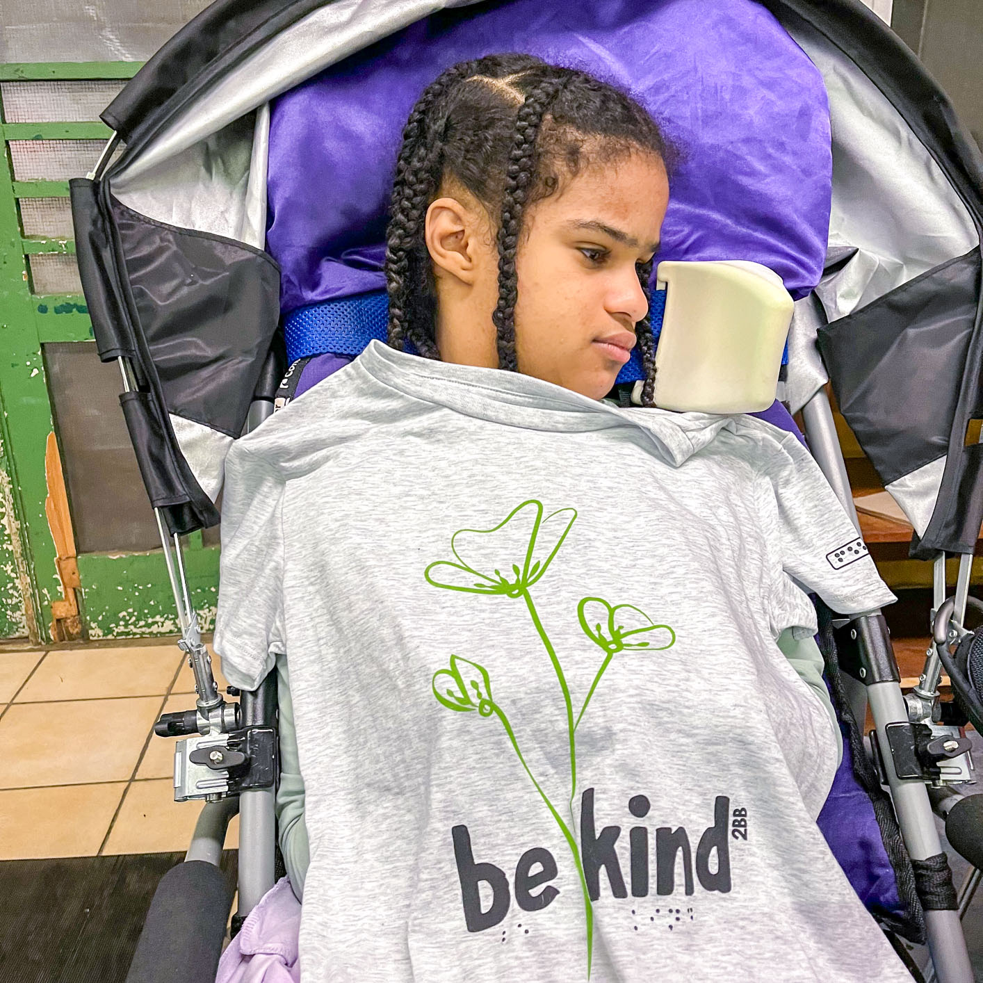 Photo of a little girl in a stroller holding a gray t-shirt from Two Blind Brothers, which has green line art of flowers with black text beneath that reads ''Be Kind'' in print and Braille letters.