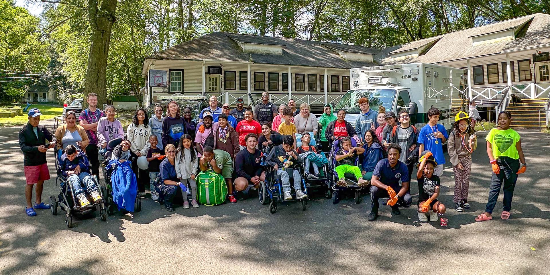 A large group photo of young children and teenagers, along with VISIONS staff all standing together by one of the cabins at VCB.