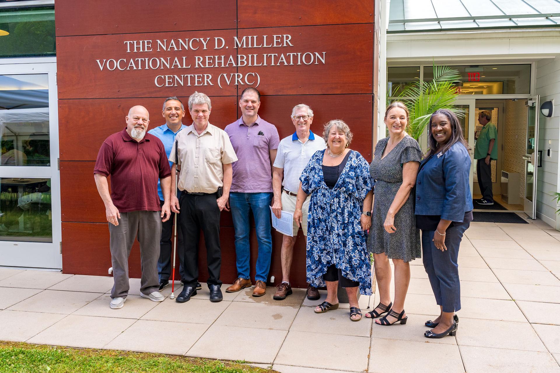 A group of VISIONS Executive Staff and Board of Directors stand outside in front of a large wall with a sign reading ''The Nancy D. Miller Vocational Rehabilitation Center'' on it.
