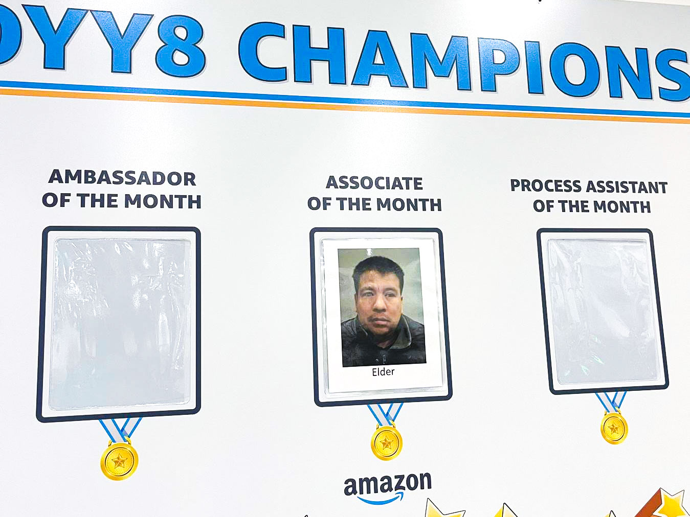 Photo of a printed photograph of a man named Elder, placed under an ''Associate of the Month'' title, on a wall titled DYY8 Champions.