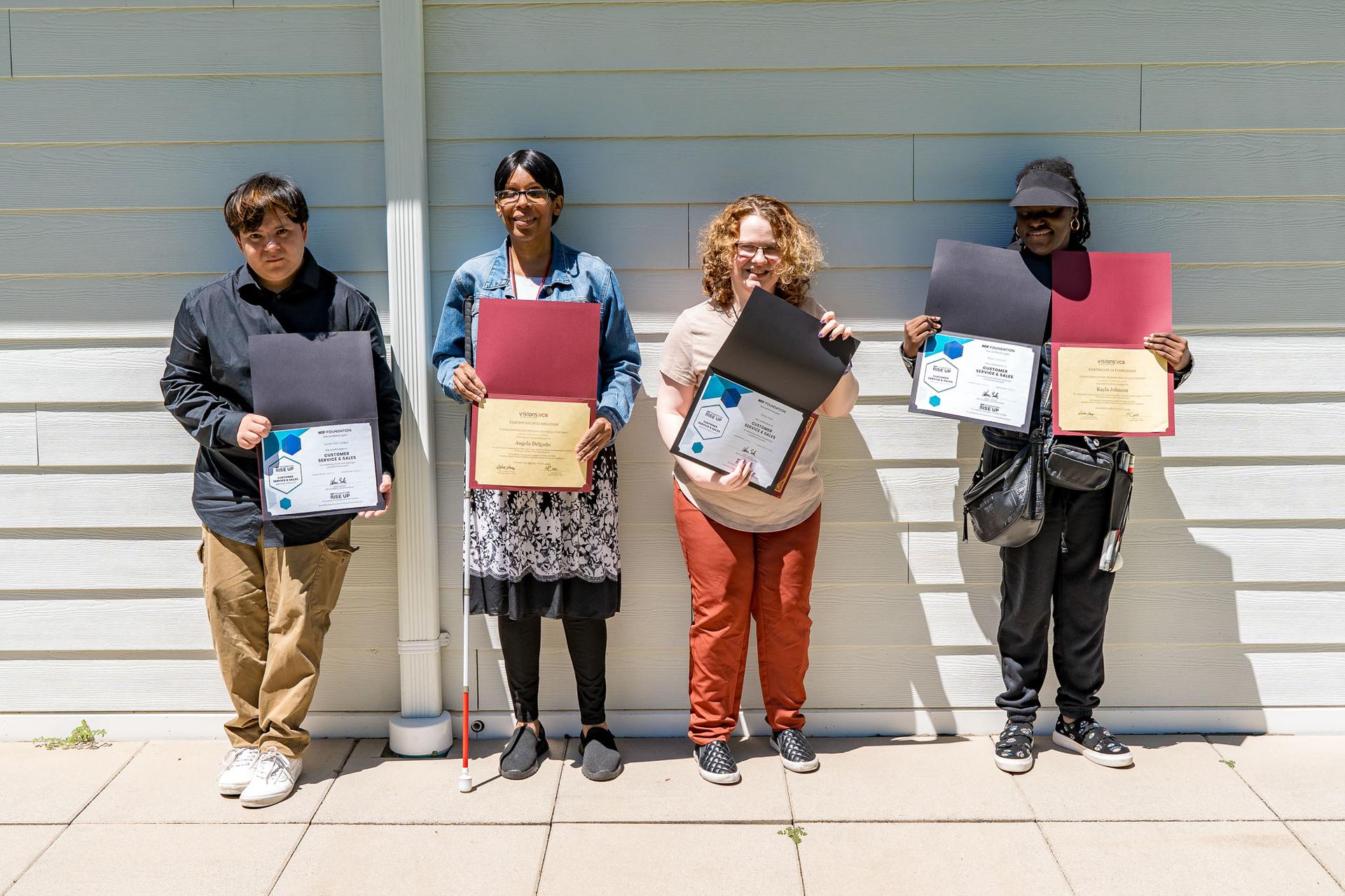 A group of four participants stand in front of a white wall and hold their certificates as they smile to the camera.