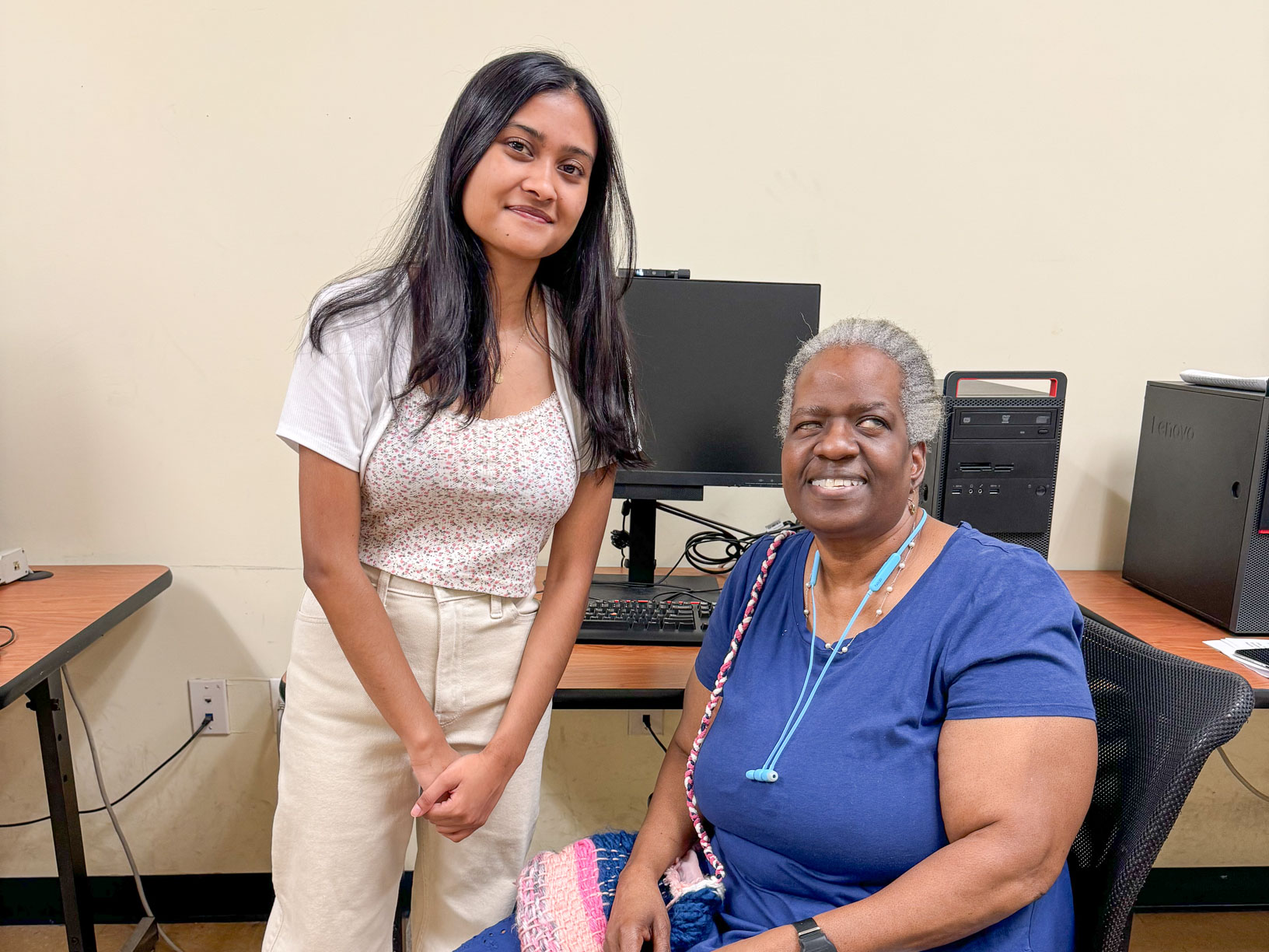 Photo of a high school-aged girl in a white dress standing beside an older adult in a blue shirt, sitting in a chair, as both smile to the camera.