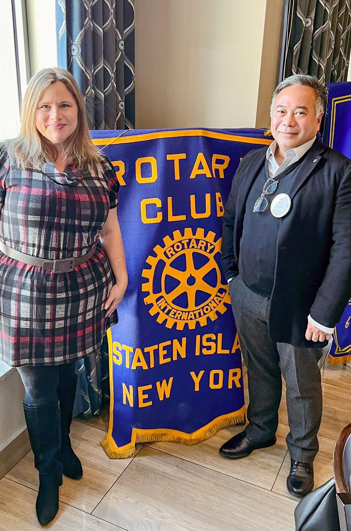 Photo of a woman and man standing and smiling to the camera, with a flag in between them. The dark blue flag with yellow text reads ''Rotary Club, Rotary International, Staten Island, New York.