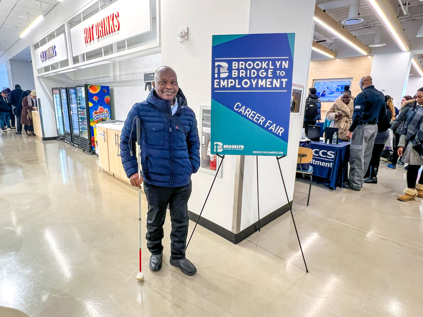 Photo of a man holding his cane, standing in front of a sign reading ''Brooklyn Bridge to Employment Career Fair''.
