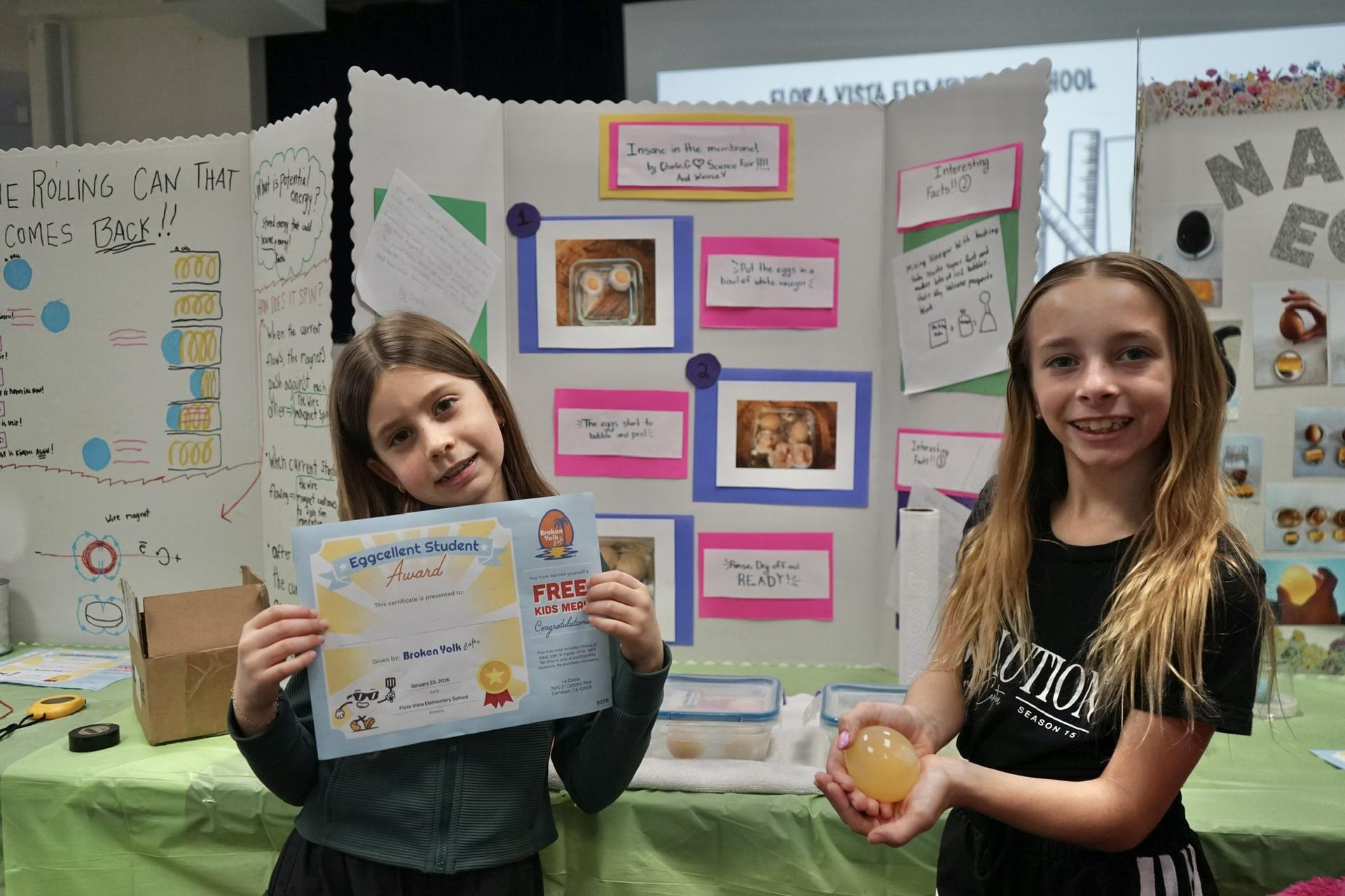 Two students stand in front of their science fair board, holding a certificate and an egg.