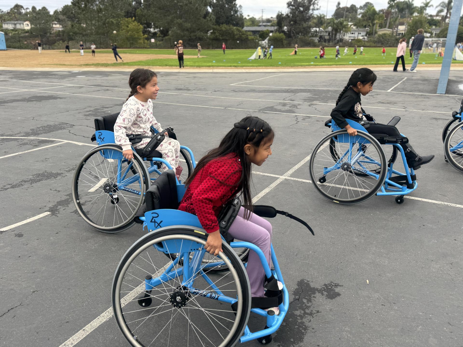 Three students try out basketball wheelchairs on the blacktop.