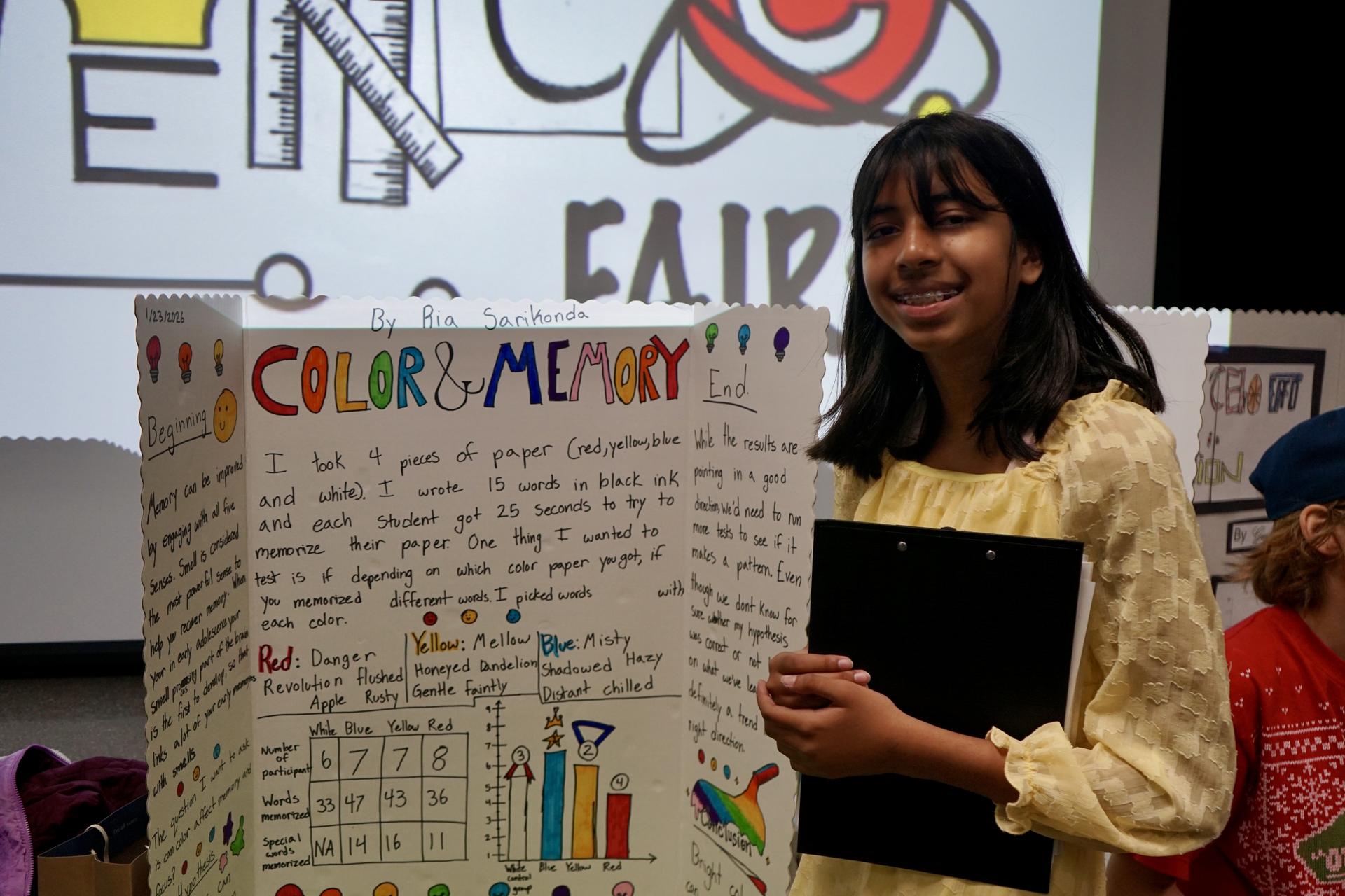A student stands in front of her science fair project smiling.