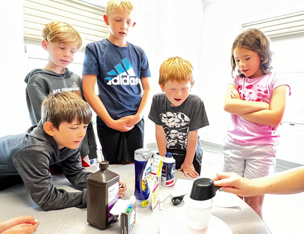 Five young kids watch science experiment on table.