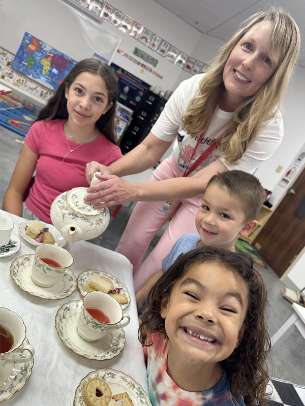 Blonde woman pouring tea into teacups for three young children.