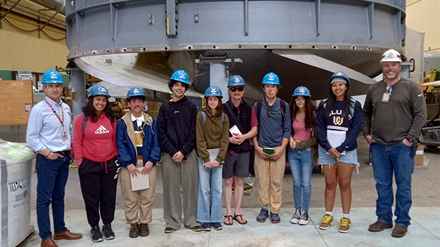 A group of people wearing hard hats stand in front of a large turbine.