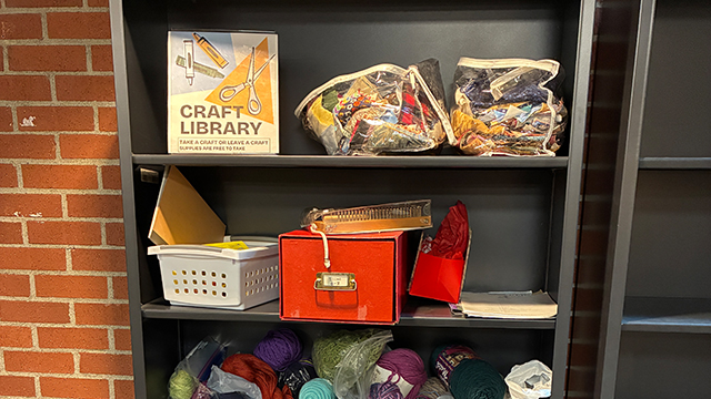 A gray bookshelf containing a sign that reads “Craft Library” and assorted craft supplies.