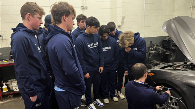 A group of students watch a technician demonstrate putting chains on car tires.