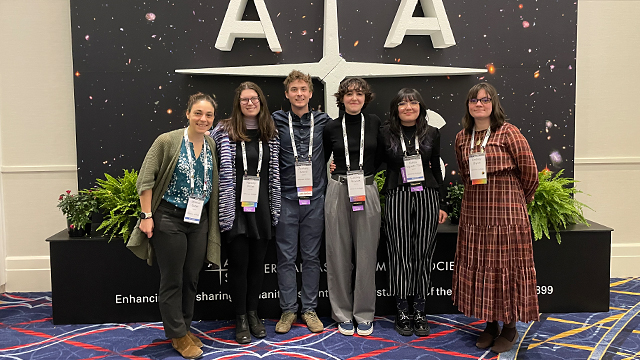 Six people wearing lanyards stand in front of a display with a large white logo on top of a background of stars in the sky.