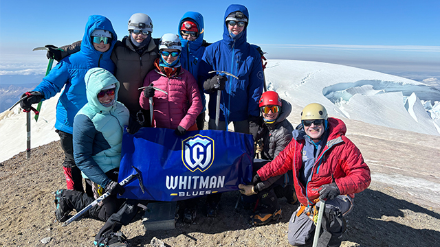 A group of climbers pose with a “Whitman Blues” banner on the top of a mountain summit.