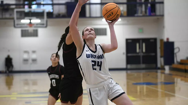 Korin Baker looks upward with a basketball in one hand. Two players from the opposing team can be seen behind her.