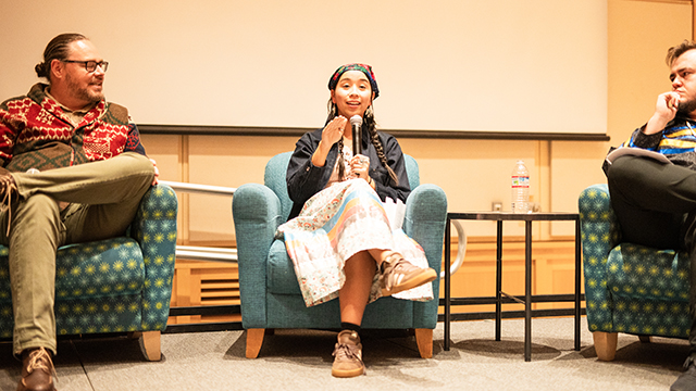 Three people seated in chairs; one holding a microphone and speaking.