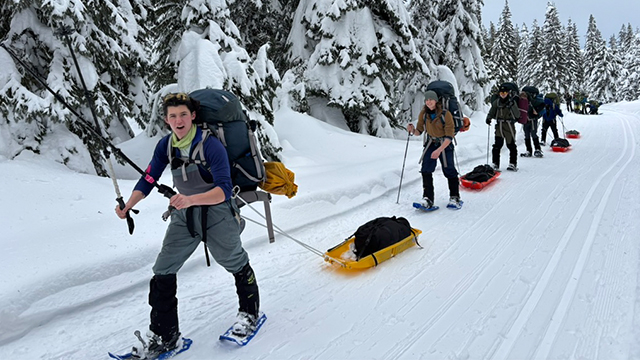 A line of people wearing snowshoes and wearing backpacks on a snowy path.