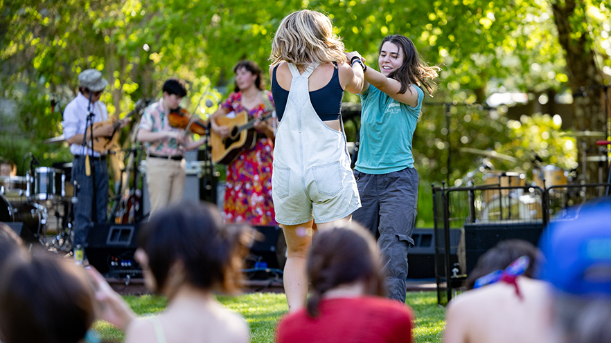 Two students dance together at Wallapalooza with several musicians visible in the background.