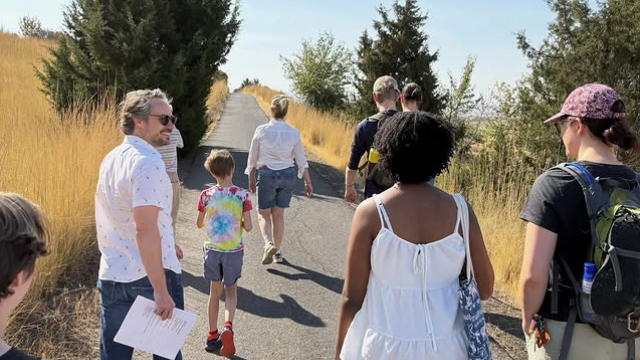 A group of people walking on a path lined with dry grass and green trees.