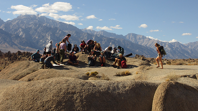 A group of people sit on rounded rocks with mountains visible in the background.