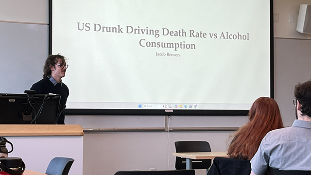 Jacob Benson '26 stands at the front of a classroom by a projection that reads “US Drunk Driving Death Rate vs Alcohol Consumption”