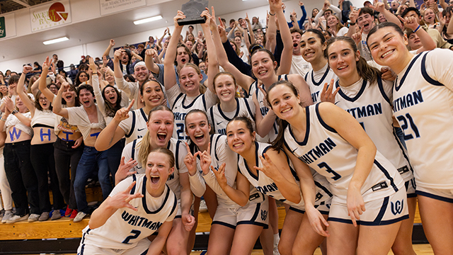 A group of Whitman women’s basketball players pose for a photo with many excited fans in the background.