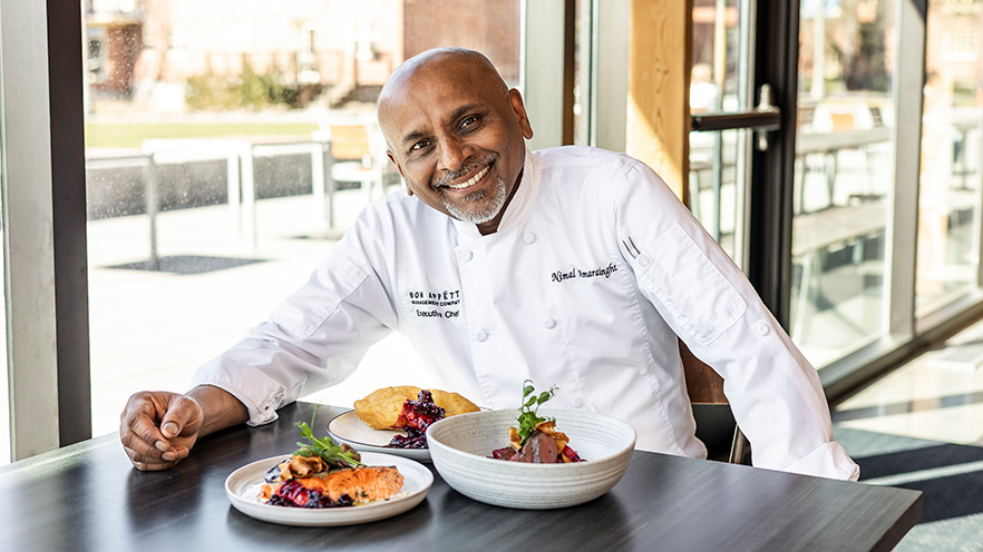 Chef Nimal Amarasinghe sits at table that contains three plates of food.