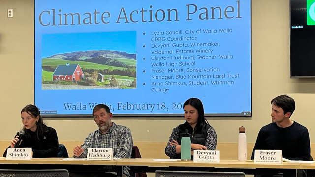 Four people sit behind a table, one holding a microphone. In the background, a presentation slide title “Climate Action Panel.”