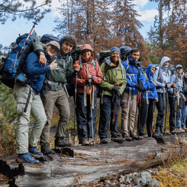 Eleven students with backpacking gear stand on a log in a meadow
