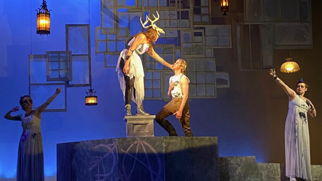 Four actors on a stage. In the center, a figure with antlers leans down from a pedestal to another actor. On the sides, two archers aim toward the central figures.